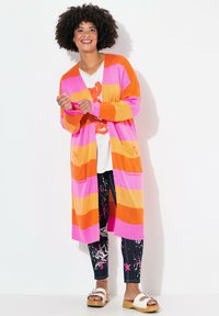 Cardigan lungo e aperto davanti con strisce orizzontali rosa, arancioni e gialle; presenta due tasche anteriori. Indossato sopra una t-shirt bianca con grafica e leggings con motivo.