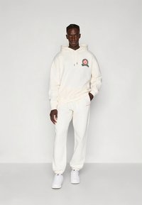 Cream-colored hoodie with embroidered logo and matching sweatpants. Made of soft fabric, featuring a relaxed fit and minimal design.