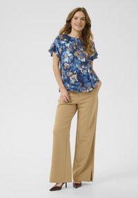Blomstret bluse i blå med korte flæseærmer, parret med brede beige bukser og burgunder hæle. Detalje med nøglehulskåret halsudskæring.