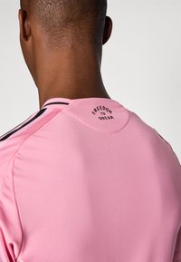 Lys pink atletisk trøje med sorte detaljer, med rund halsudskæring og teksten "FRIHED TIL AT DRØMME" på bagsiden af halsudskæringen. Glat tekstur.