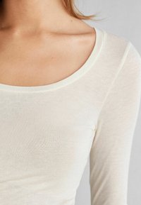 Langärmliges Oberteil in Creme mit weitem, rundem Ausschnitt und weichem, leichtem Stoff. Glatte Textur, minimales Design ohne Muster.