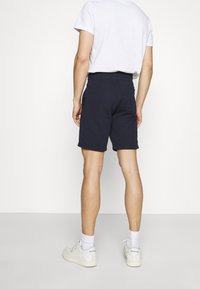 Homme de dos portant un t-shirt blanc, un short bleu marine, des chaussettes blanches et des baskets Reebok blanches, sur fond uni.