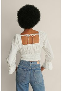 Kobieta z kręconymi włosami, odwrócona plecami, ubrana w białą bluzkę z długim rękawem, marszczoną, z odkrytymi plecami oraz niebieskie dżinsy z wysokim stanem.