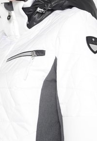 Veste matelassée blanche avec col noir, poche poitrine zippée, panneau latéral gris et patch de marque noir et argent sur la manche supérieure.