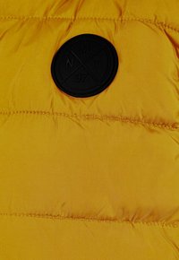 Gul vadderad jacka med en quiltad textur och en svart cirkulär logotyp-patch med initialer och ett korsat mönster.