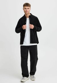 Zwarte corduroy overhemd en broek, gecombineerd met een wit t-shirt. Opvallende details zijn een zak en knoopsluiting op het shirt, casual sneakers.