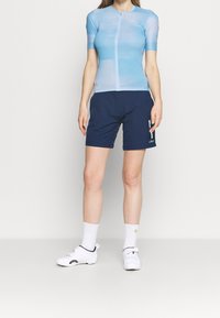 Blå kortärmad cykeltröja med dragkedja framtill, tillsammans med marindrabbade shorts med en logotyp. Vita cykelskor och strumpor.