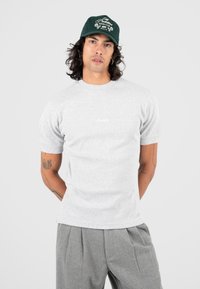 Uomo con capelli lunghi che indossa un berretto verde, una maglietta grigia chiara a maniche corte e pantaloni grigi plissettati su uno sfondo neutro.