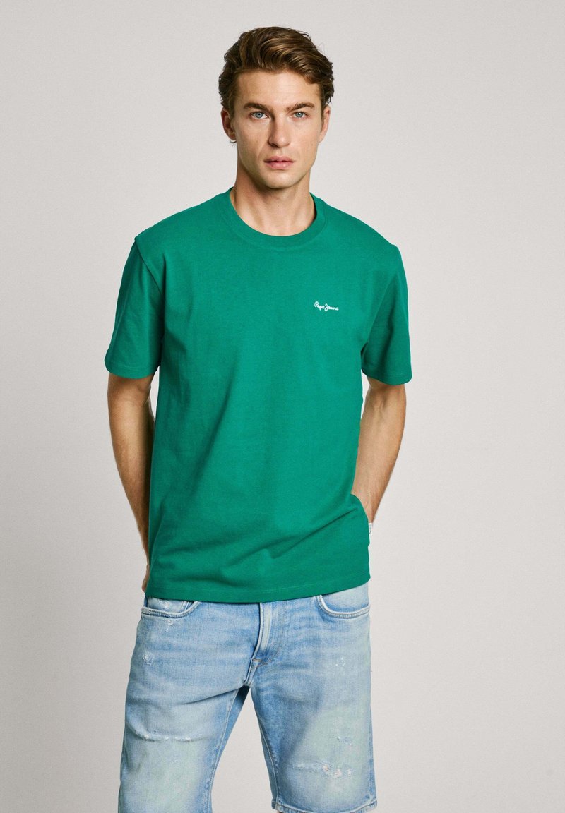 Green cotton t-shirt with a crew neckline and short sleeves, featuring a small white logo on the left chest. Worn with light denim shorts.