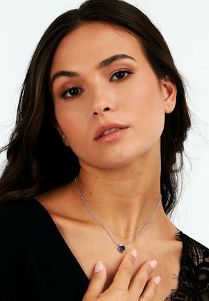 Silberne Halskette mit einem herzförmigen blauen Edelstein-Anhänger, getragen von einer Person mit langen dunklen Haaren und einem schwarzen Oberteil mit Spitzenbesatz.