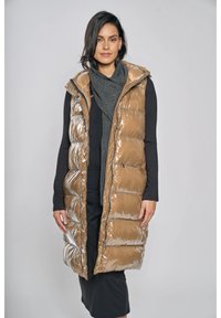 Lange, gewatteerde bodywarmer in glanzend beige materiaal met een capuchon, voorzien van gestikte secties en zijzakken, gedragen over een donkere outfit.