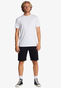 T-shirt bianco di cotone con maniche corte abbinato a pantaloni shorts neri. Il modello indossa sneaker nere con lacci bianchi e calzini bianchi.