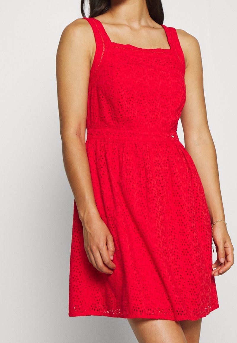 Femme portant une robe rouge sans manches en œillets, avec un décolleté carré et une taille cintrée, debout devant un fond gris clair uni.