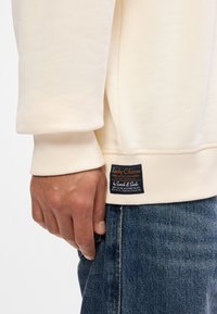 Κρέμα φούτερ με ριμπ μανσέτες, που φέρει μια μπλε και πορτοκαλί ετικέτα μάρκας κοντά στο στρίφωμα. Φ worn με τζιν παντελόνι.