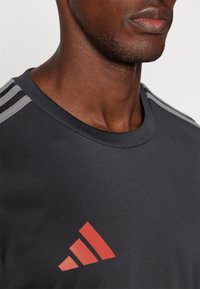 Schwarzes Sport-T-Shirt mit grauen Streifen an den Schultern und rotem diagonalen Logo. Glattes Material mit Rundhalsauschnitt. Fokus auf dem oberen Brustbereich.