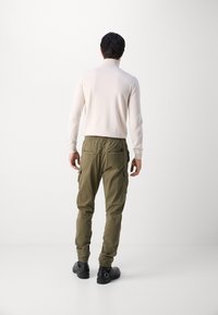 Helles beige Rollkragenpullover, schmal geschnitten mit langen Ärmeln, kombiniert mit olivgrünen Cargo-Shorts mit Seitentaschen und elastischen Bündchen. Schwarze Schuhe.