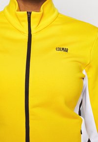 Żółta kurtka zapinana na zamek z wysokim kołnierzem. Czarny pionowy zamek błyskawiczny i akcenty wzdłuż boków. Logo "COLMAR" w kolorze czarnym na klatce piersiowej.