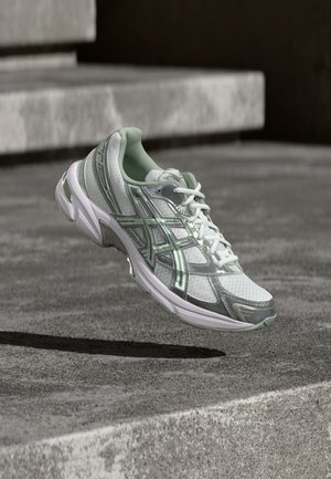 Adidași albi și argintii cu accente verde menta, suspendați în aer deasupra unei suprafețe de beton gri, cu trepte în fundal.