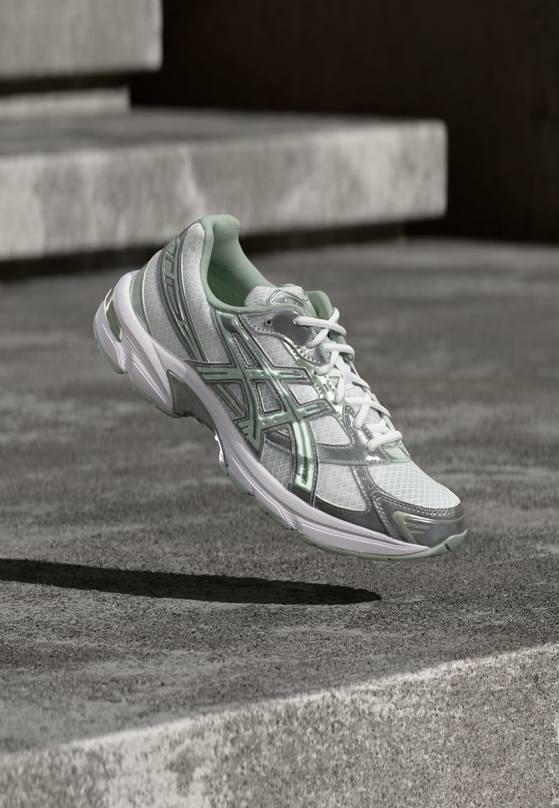 Λευκό και ασημένιο αθλητικό παπούτσι με μαντζέντα πράσινες λεπτομέρειες, αιωρούμενο στον αέρα πάνω από μια γκρι τσιμεντένια επιφάνεια με σκαλοπάτια στο φόντο.