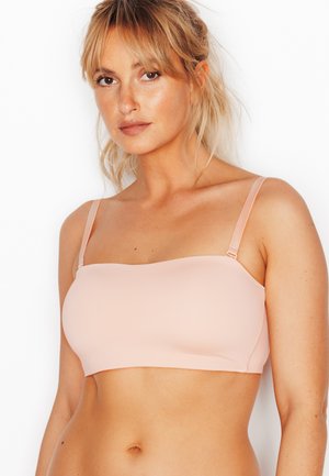 Giovane donna con capelli biondi che indossa un reggiseno a fascia rosa chiaro con sottili spalline regolabili su sfondo bianco.