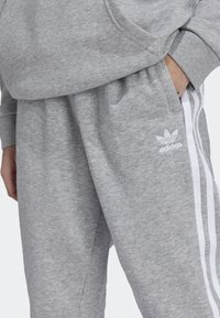 Pantalones de chándal grises hechos de una tela suave con un corte recto, que presentan rayas laterales blancas y un logo de Adidas en la izquierda.