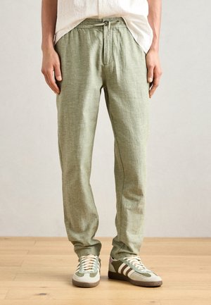 Pantalon vert clair près du corps avec taille à cordon, porté avec des baskets à lacets blanches et vertes sur un sol en bois.