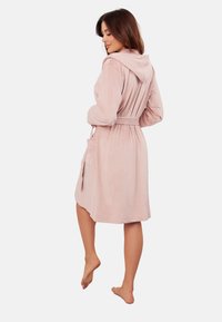 Jasnoróżowy szlafrok z kapturem, wykonany z miękkiej tkaniny, z paskiem do związania i bocznymi kieszeniami, posiadający długie rękawy i luźny krój.