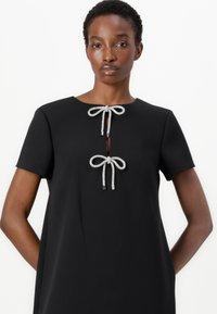 Abito nero con maniche corte, caratterizzato da due fiocchi di perline argento sulla scollatura, taglio verticale e una texture del tessuto liscia.