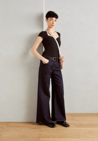 Combinaison côtelée noire avec un décolleté en V profond, associée à un jean en denim foncé à jambe large. Mise en valeur par une ceinture noire et un sac à bandoulière beige.