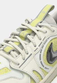 Nike tenisice s bijelom kožom, sivim naglascima, mrežastim panelima u svijetložutoj boji, sjajnim crnim logotipom i teksturiranim vezicama na podstavljenom jeziku.