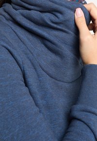 Modrá svetr s kapucí vyrobená z texturované pleteniny. Má žebrované manžety a volný výstřih, který ukazuje měkký, teplý materiál.