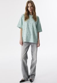 Mátově zelené oversized tričko s malými černými tečkami, v kombinaci se světle šedými džíny se stříbrnými ozdobami, doplněné černými špičatými balerínami.