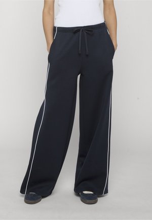 Osoba nosząca granatowe spodnie z szerokimi nogawkami i białymi paskami po bokach, ze ściągaczem w talii, ręce w kieszeniach oraz ciemne trampki na jednolitym tle.