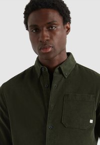 Camicia in velluto a coste verde con bottoni e taschino sul petto, caratterizzata da un tessuto testurizzato e colletto. Il design è di vestibilità standard con dettagli classici.
