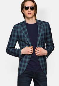 Tkaninowa marynarka w kratę w ciemnozielonym i granatowym kolorze, z zapięciem na jeden guzik i klapami, zestawiona z granatowym t-shirtem i ciemnymi dżinsami.