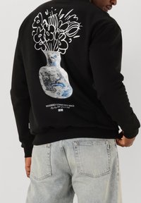 Svart sweatshirt med en stor grafisk avbildning av en blå och vit vas med stiliserade blommor på baksidan, med text under bilden.