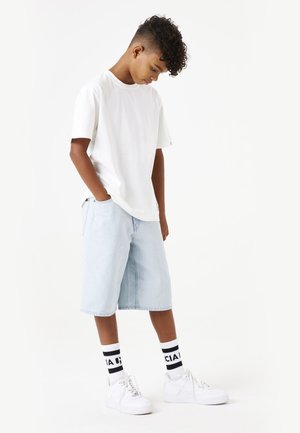 Camiseta blanca de manga corta, pantalones cortos de mezclilla azul claro, calcetines a rayas en blanco y negro, y zapatillas blancas. Estilo de ropa de calle informal.