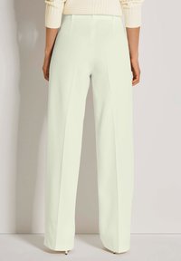 Vrouw in hoge taille, wijde witte broek en een crèmekleurige gebreide trui, staande tegen een effen lichte muur.