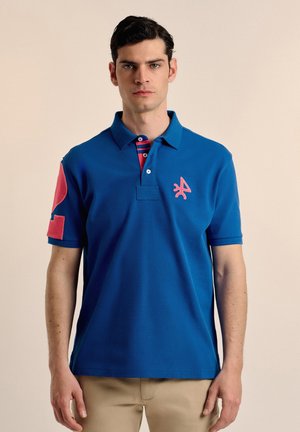 Camisa polo azul hecha de una mezcla de algodón, con un logotipo rosa en el pecho y detalles rosas en las mangas, con un cuello a rayas.