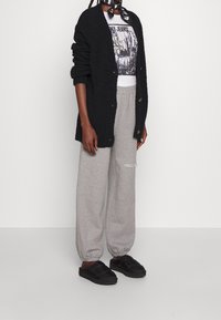 Person, der einen schwarzen Cardigan, ein grafisches weißes T-Shirt, graue Jogginghosen mit einem Riss, schwarze Hausschuhe und einen schwarz-weißen Schal trägt.