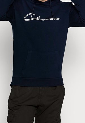 Persoon draagt een marineblauwe hoodie met een wit scriptlogo en zwarte cargobroek, armen omhoog, tegen een effen lichte achtergrond.