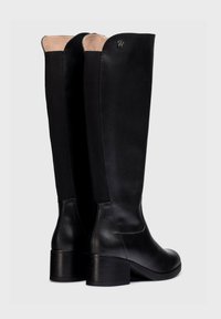 Botas de cuero negras hasta la rodilla con tacón cuadrado, con un acabado suave, forro beige en contraste y un pequeño logo dorado en la parte superior.