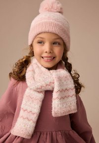 Rosa gestrickte Mütze mit pompon und passendem flauschigen Schal mit Zickzackmustern und goldenen Akzenten. Getragen über einem rosa strukturierten Kleid.