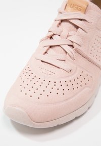 Růžové semišové tenisky s perforovaným svrškem, plochými tkaničkami a texturovanou gumovou podrážkou. Obsahují malé logo UGG na jazyku.