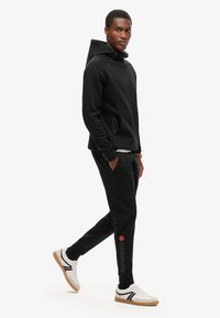 Zwarte hoodie met een kangoeroezak en verstelbare capuchon, gecombineerd met zwarte taps toelopende joggingsbroek met een oranje logo en witte sneakers.
