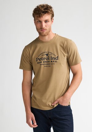 Mężczyzna o kręconych włosach, ubrany w brązowy t-shirt Petrol Ind i ciemne jeansy, stoi z jedną ręką w kieszeni na jednolitym tle.
