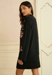 Fekete pulóverruha színes virágos hímzéssel a vállakon. Rázott mandzsetták és szegély, laza szabással és puha textúrával.