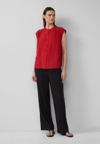Chemise rouge à manches courtes avec un col rond, devant à boutons et détail froncé aux épaules, associée à un pantalon noir ample et des sandales.