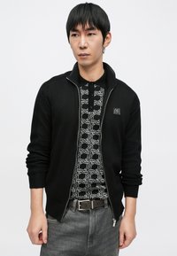 Juodas megztinis su užtrauktuku ir ribinėmis rankogaliais, dėvimas ant juodos ir baltos raštuotos polo marškinėlių. Pilki džinsai ir ruda diržas užbaigia aprangą.