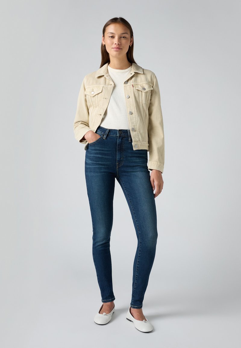 Beżowa kurtka dżinsowa, biała koszulka, ciemnoniebieskie skinny jeans, płaskie białe buty. Prosty design z zapięciem na guzik i przednimi kieszeniami.
